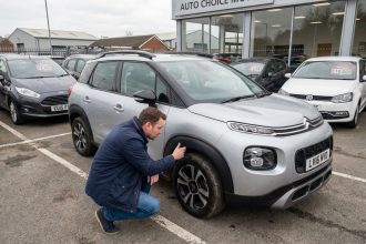 Un homme inspecte attentivement la carrosserie d'une Citroën C3 Aircross d'occasion dans un parking de concessionnaire automobile